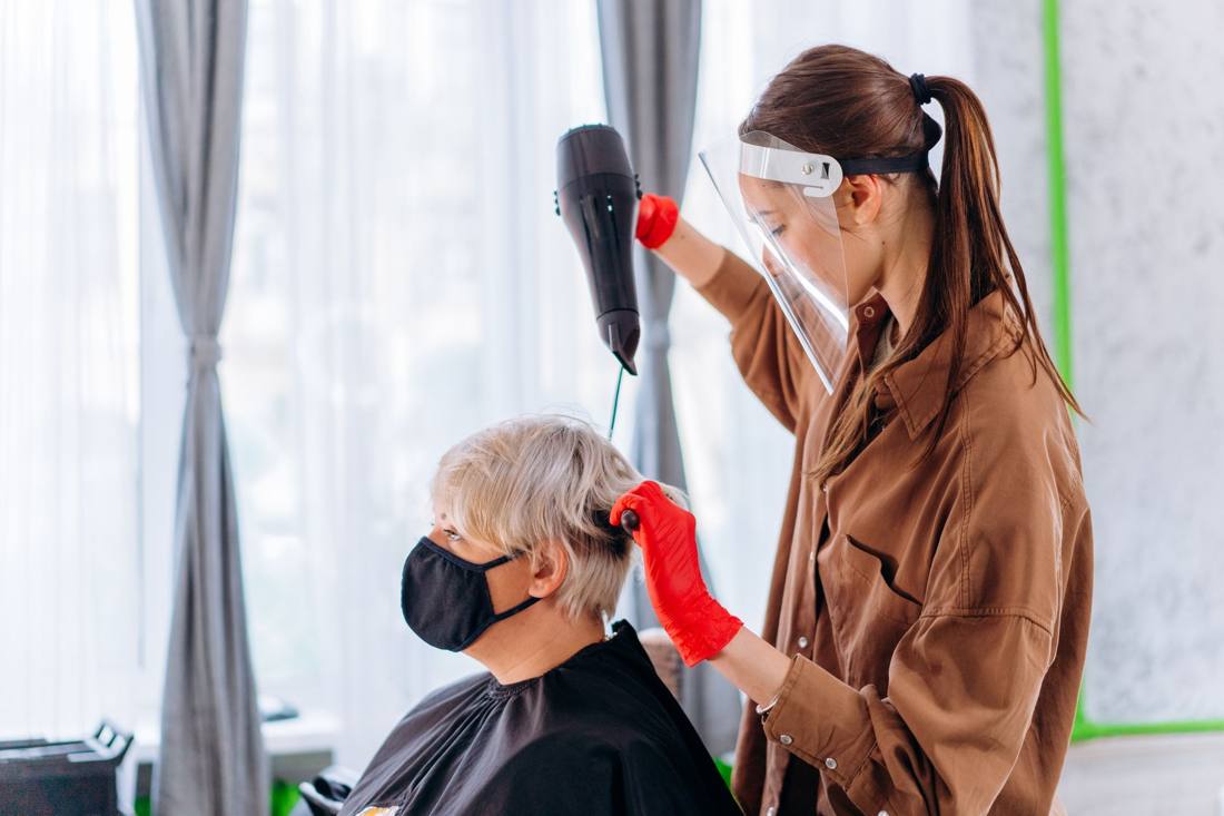 A hair stylist wearing a face shield and gloves dries a client's hair in a salon setting.